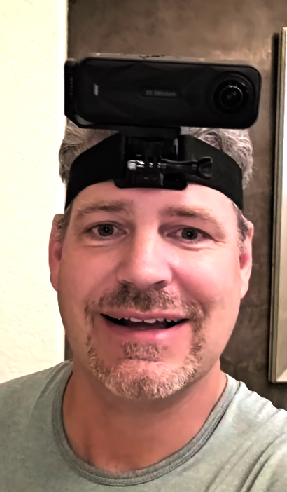 A contributor photographed indoors wearing a compact dual-lens 360 camera clipped to a quick-release buckle on an elastic head strap, positioned above the forehead.